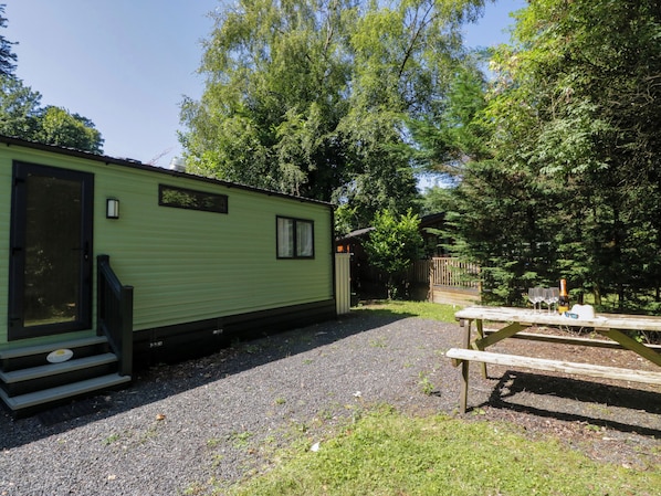Una casa móvil verde con un porche y escalones, rodeada de árboles y una mesa de picnic.
