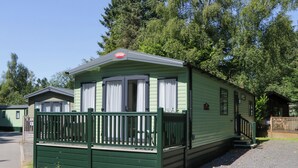 A green mobile home with a porch and a small deck, surrounded by trees. - High Perch (Windermere)