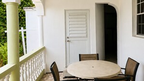 Uma área de pátio coberta com uma mesa redonda e quatro cadeiras, uma porta branca e uma janela com vista para o verde.