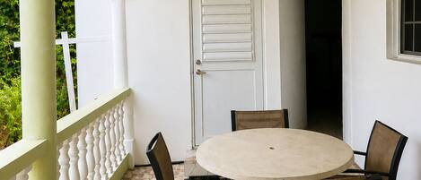 Un porche cubierto con una mesa redonda y cuatro sillas, una puerta blanca y una ventana con vistas a zonas verdes.