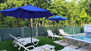Zona junto a la piscina con césped artificial, sillas reclinables blancas y sombrillas azules.