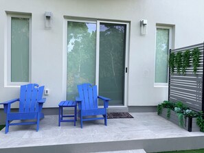 Eine Terrasse mit zwei blauen Stühlen, einem kleinen Tisch, einer Schiebetür und einem Pflanzgefäß mit Grünpflanzen.