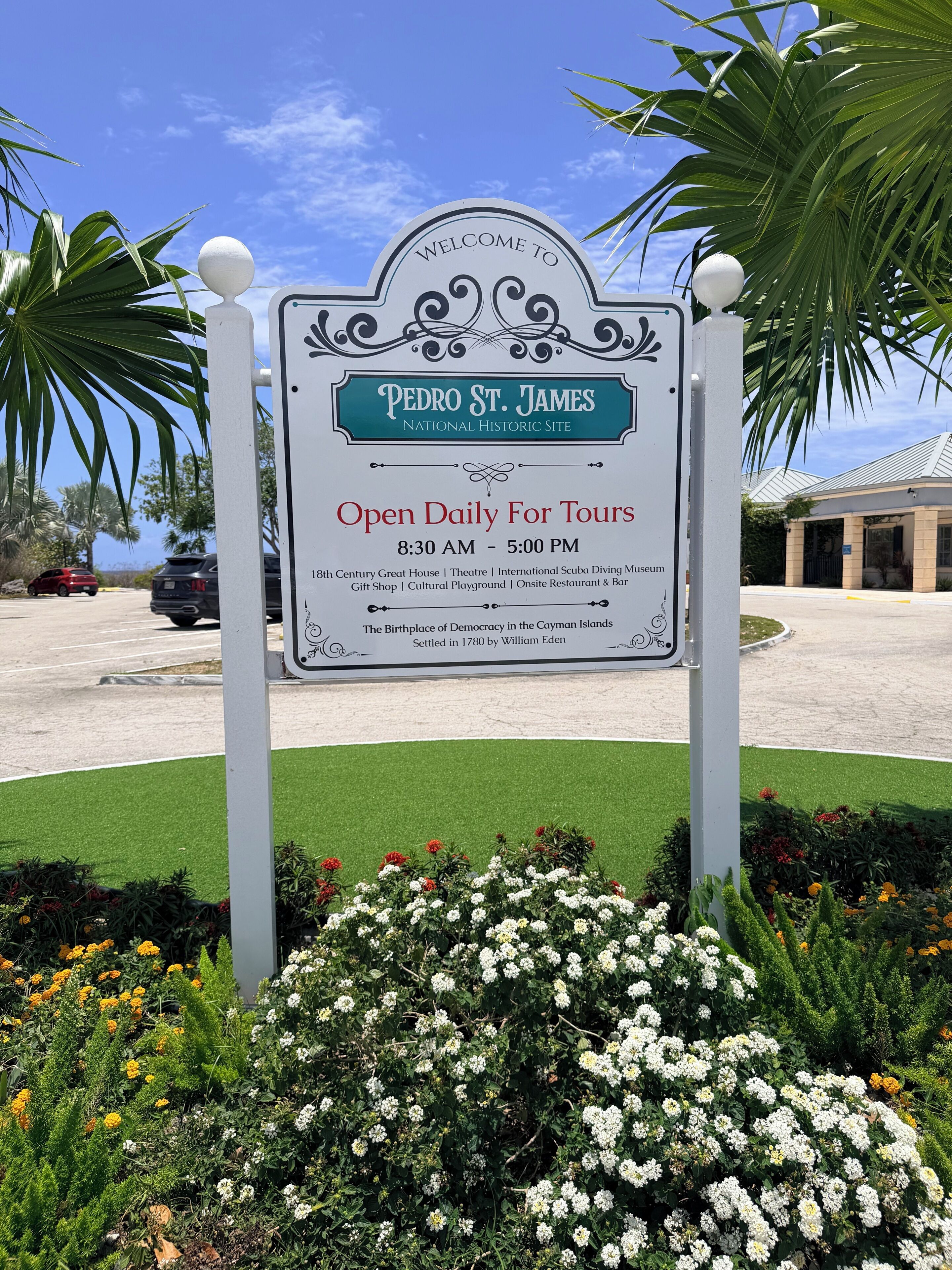 A sign for the Pedro St. James National Historic Site, open daily for tours from 8:30 AM to 5:00 PM, featuring the 18th Century Great House, Theatre, International Scuba Diving Museum, Gift Shop, Cultural Playground, and Onsite Restaurant & Bar.