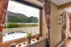 A room with a large window overlooking a lake and mountains, a hot tub, and a wooden interior.