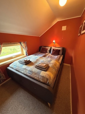 A bedroom with a bed, a window with curtains, a wall-mounted lamp, and a carpeted floor.