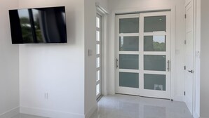 En modern inredning med en platt-TV som är monterad på väggen, en skjutdörr i glas som leder ut till ett utomhusområde och ett stilrent, kaklat golv.