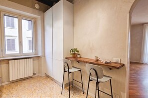 Eine kleine Küche mit einer Holzausziehtheke, zwei Barhocker, einem Heizkörper und einem Fenster mit Rollläden.