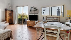 Een ruime woonkamer met een groot raam, een houten kast, een eettafel met stoelen en een televisie.