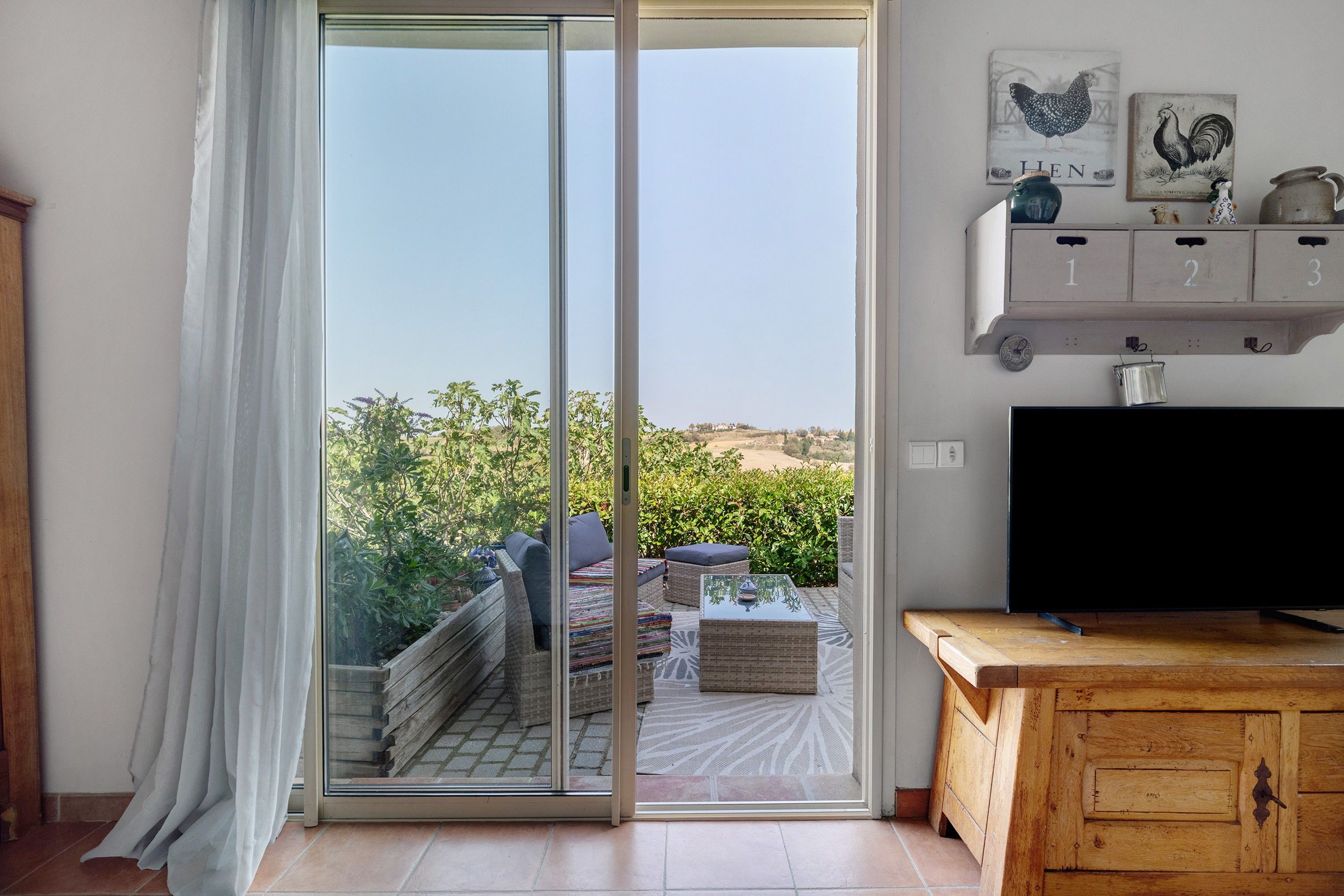 Een kamer met een schuifglasdeur die uitkomt op een balkon, een televisie op een houten kast en een plank met genummerde opbergdozen.