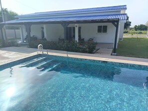 A swimming pool with a tiled edge, a covered patio area, and a building with a blue roof in the background. - Quiet 2-Bedroom House with Pool · Ideal for Long Stays (Punta Chame)