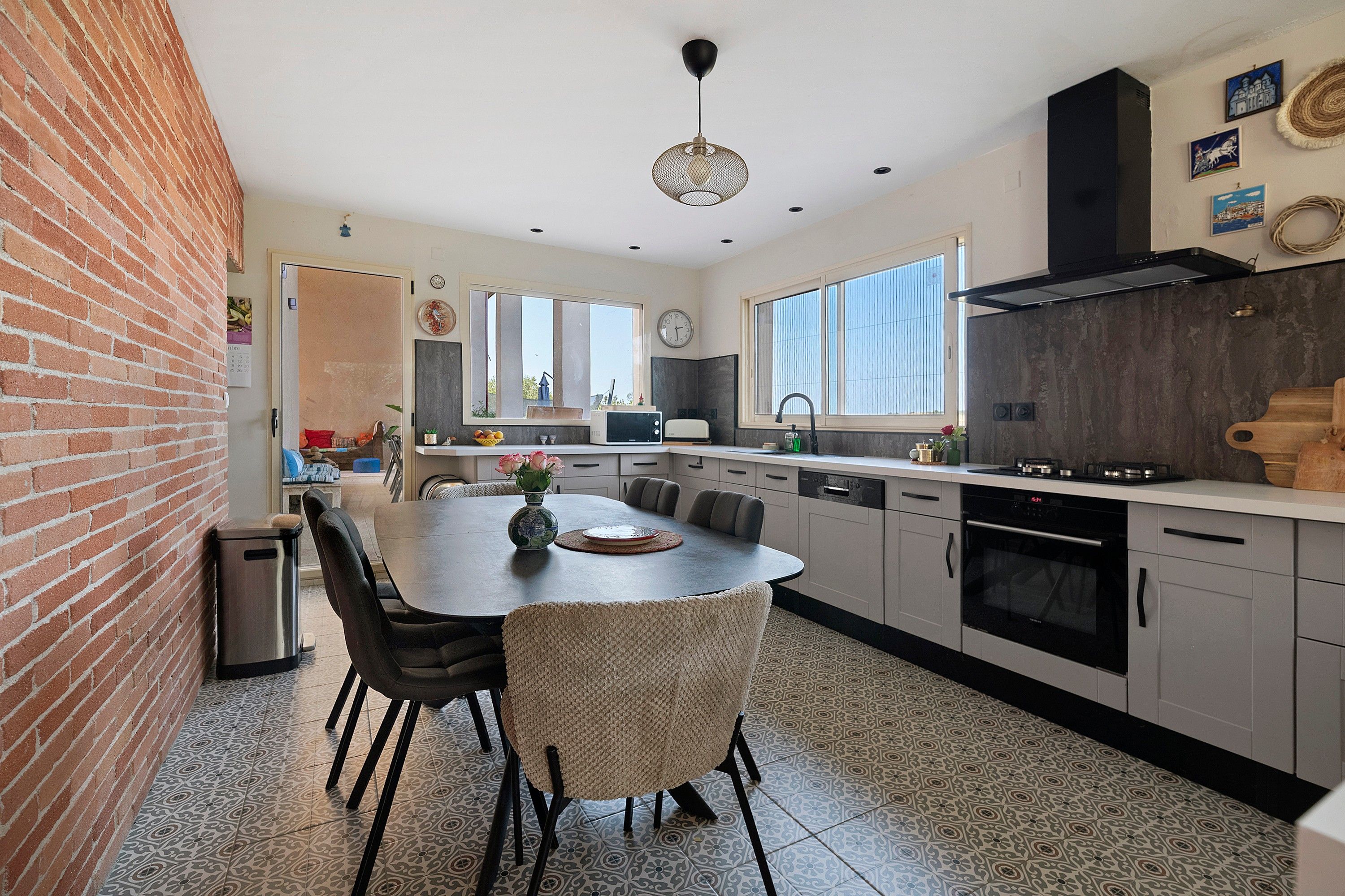 Een moderne keuken met een centraal eiland, eettafels met stoelen, een inbouwoven en een groot raam dat voor natuurlijk licht zorgt.