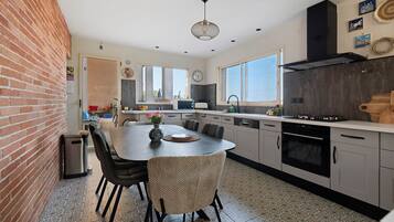 Een moderne keuken met een centraal eiland, eettafels met stoelen, een inbouwoven en een groot raam dat voor natuurlijk licht zorgt.