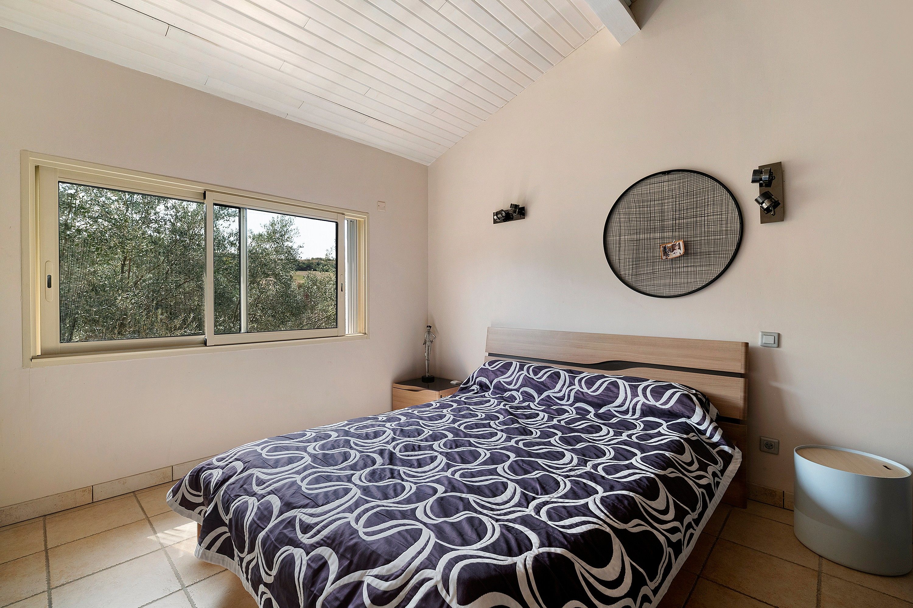 Un dormitorio con cama, ventana, lámpara de pared y un cubo de basura.
