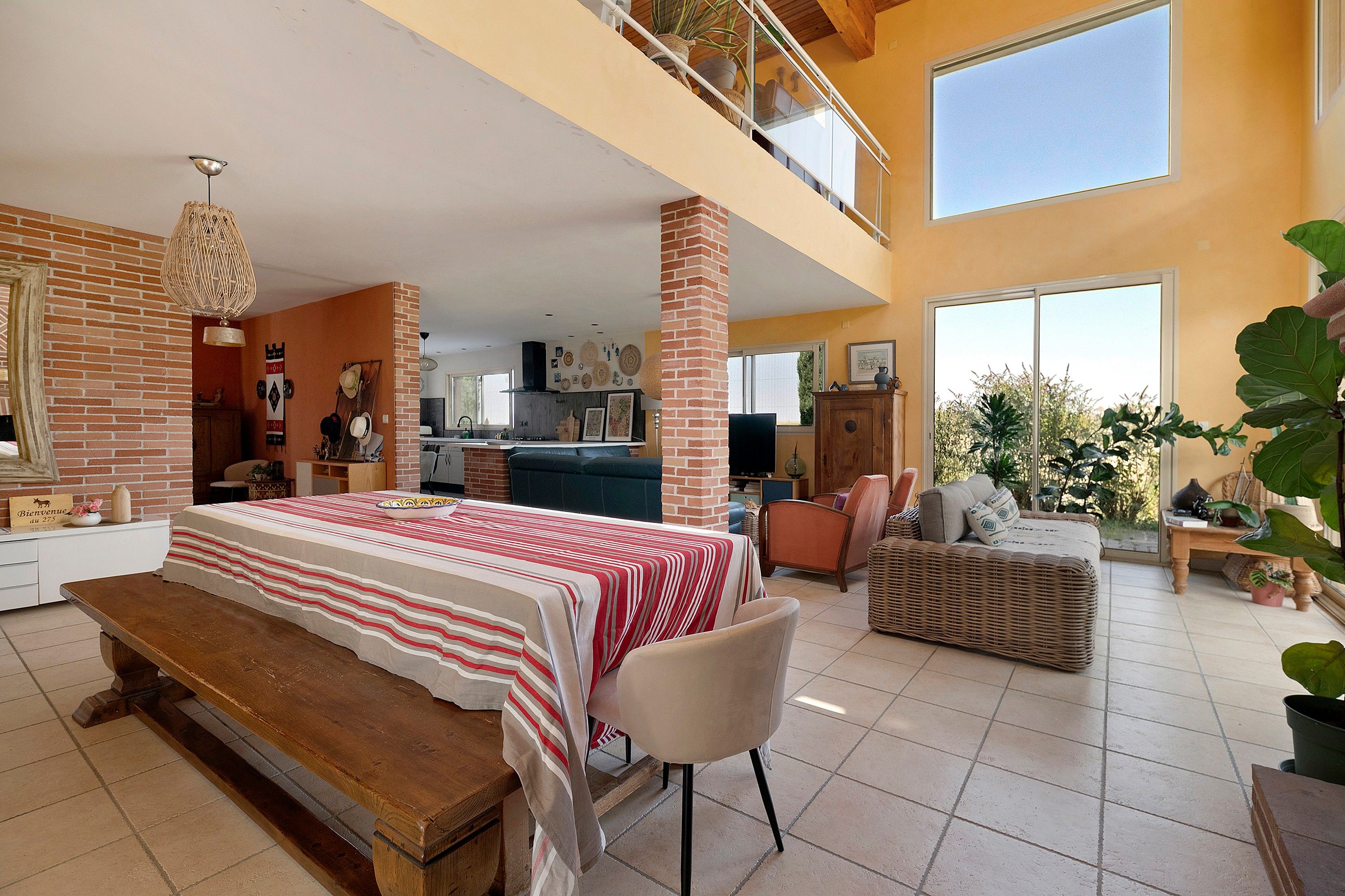 A spacious living area with a high ceiling, a balcony, and a kitchen in the background.