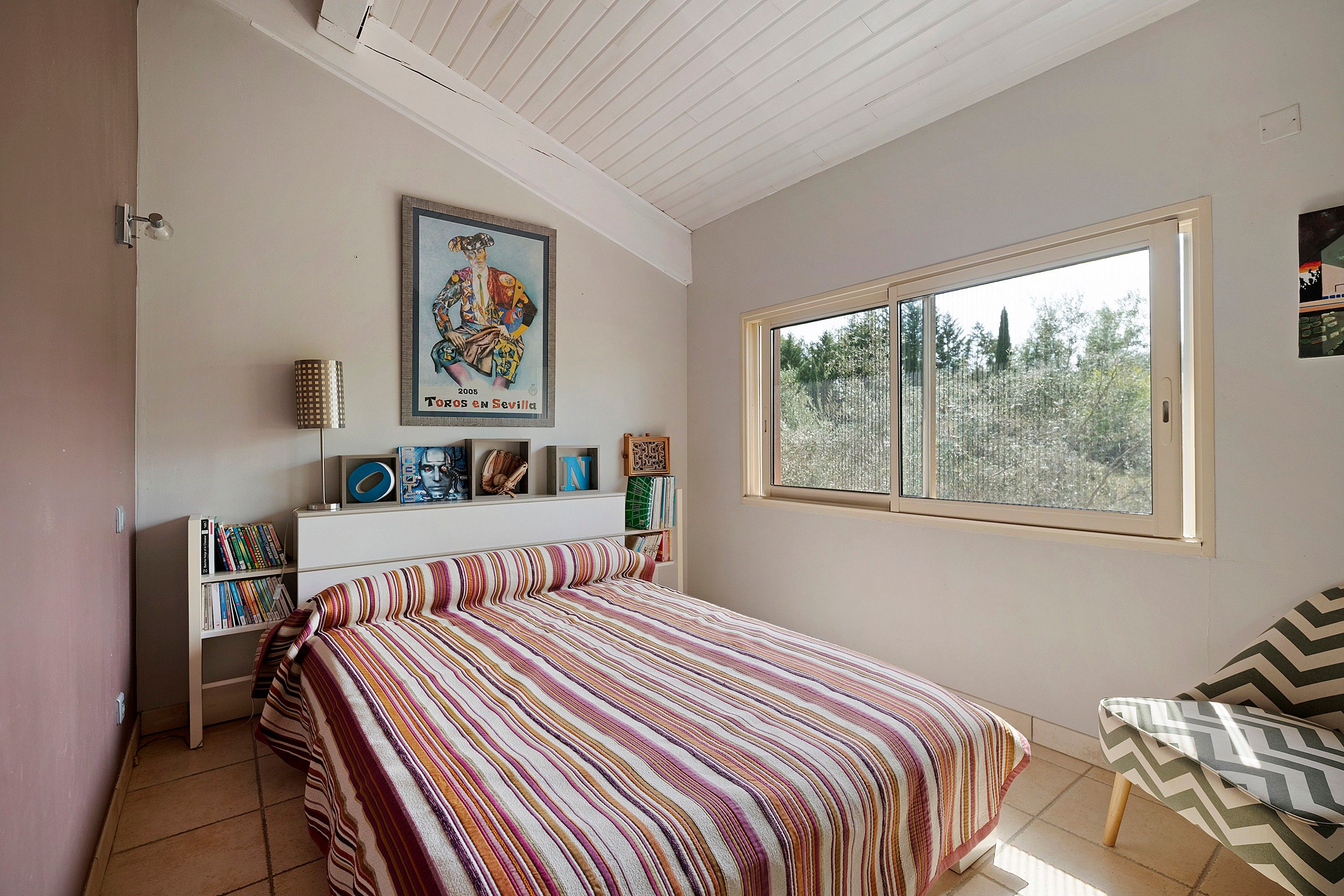 Une chambre à coucher avec un lit, une fenêtre donnant sur des arbres, une bibliothèque et un tableau encadré accroché au mur.