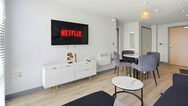 Ein modernes Wohnzimmer mit einem Flachbildfernseher, der 'NETFLIX' zeigt, einer weißen Konsole, einem runden Couchtisch und einem Essbereich mit Stühlen.
