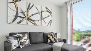 Ein modernes Wohnzimmer mit einer schwarzen Couch, abstrakter Wandkunst und Blick durch ein Fenster mit Jalousien.
