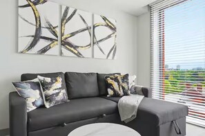 A modern living room with a black sofa, abstract wall art, and a view through a window with blinds. - Apartment Sleeps 4 - Air Con, City Centre, Wifi (Birmingham)