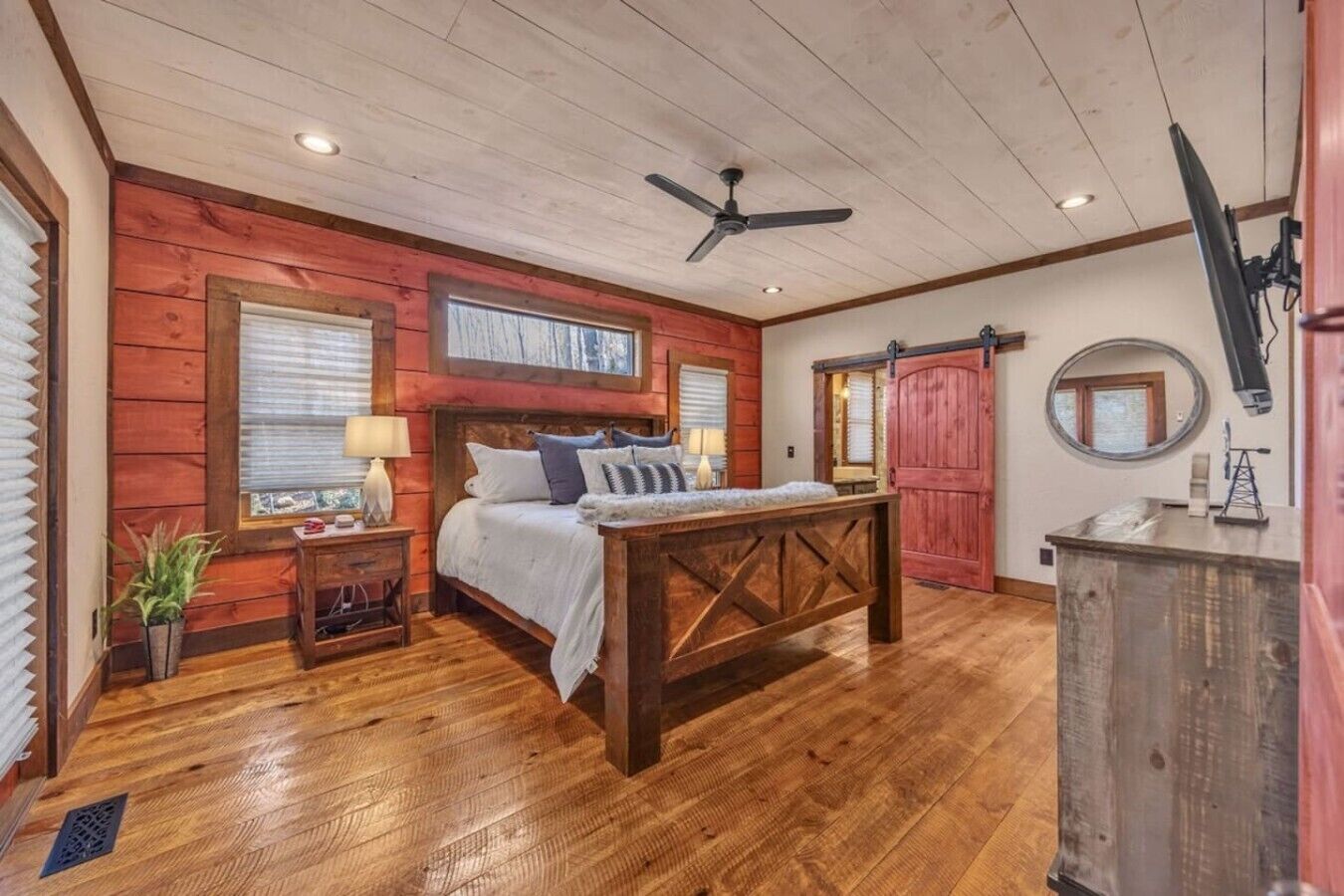 A bedroom with a wooden ceiling, a bed with pillows, a nightstand, a desk, and a red door.