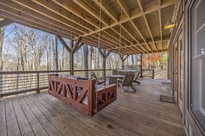 A covered outdoor deck with a wooden floor, a hanging swing, and a grill area. - Blue Ridge Escape | Luxury Mtn. Cabin near Lake BR (Blue Ridge)