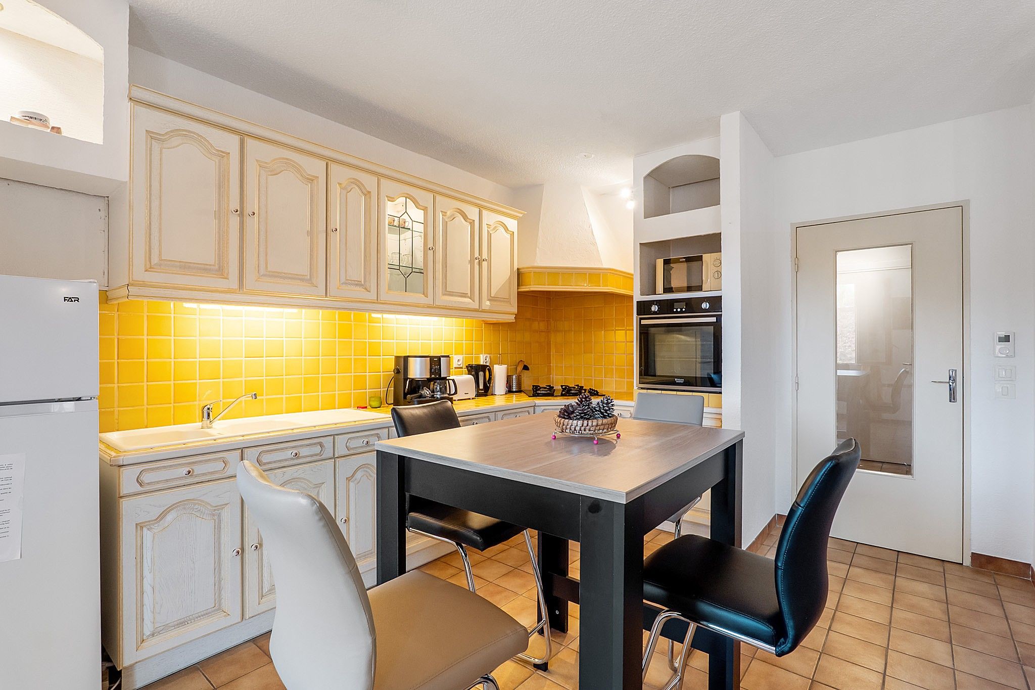 Een keuken met witte kasten, een gele achterwand, een eettafel met stoelen en een deur met een handgreep.