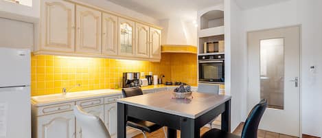 Une cuisine avec des meubles blancs, un pare-feu jaune, une table à manger avec des chaises et une porte dotée d’une poignée.