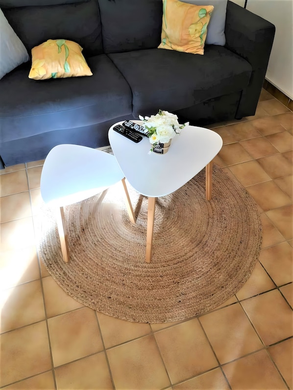 A living room with a sofa, two white chairs, a woven rug, and a small table with a flower vase and remote controls.