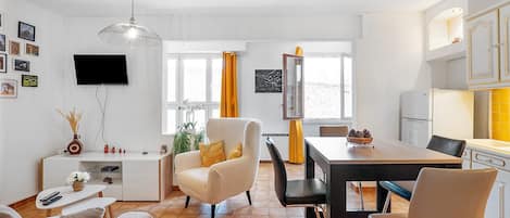 A modern living room with a television, sofa, dining table, chairs, and a kitchen area with cabinets and a refrigerator.