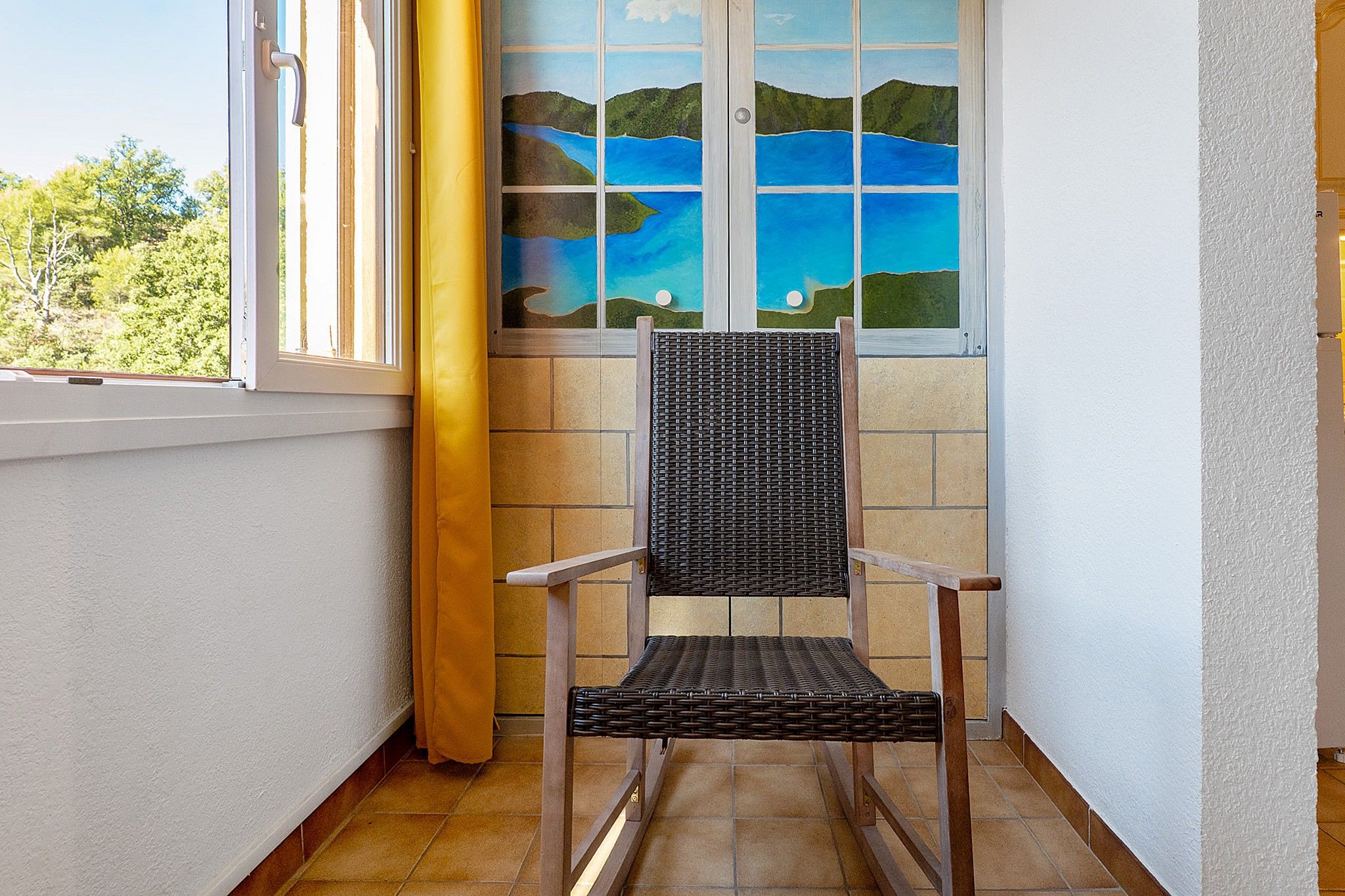 Une pièce d’angle avec un fauteuil en osier, une fenêtre donnant sur une étendue d’eau bleue et des collines verdoyantes, et un rideau jaune.