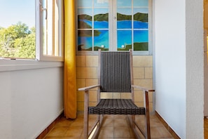 A corner room with a wicker chair, a window with a view of a blue body of water and green hills, and a yellow curtain. - Chez Géraldine Apartment (Sainte-Croix-du-Verdon)