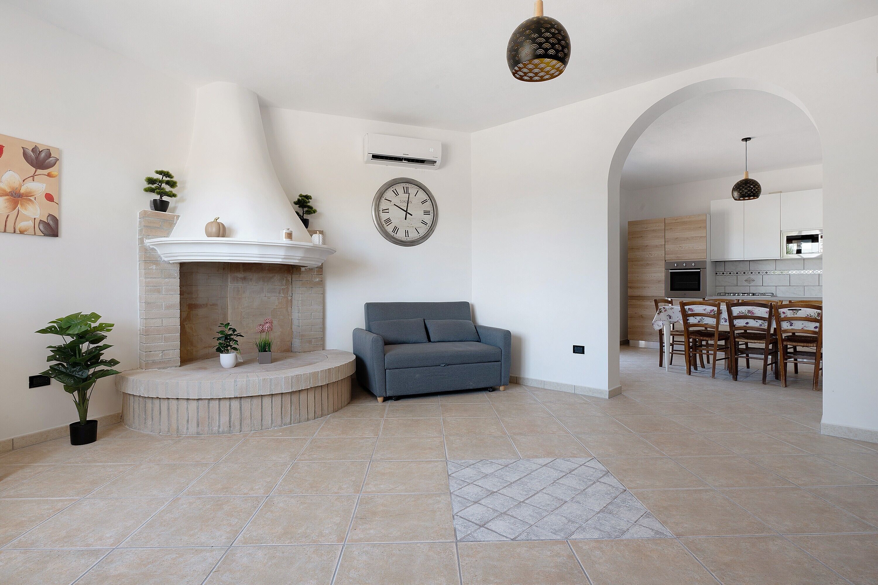 A modern living room with a fireplace, a sofa, a dining area with wooden chairs, and a kitchen in the background.