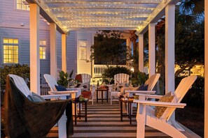 A covered outdoor seating area with white chairs, a table with a fruit basket, and string lights. - Suncerre- 100 Steps to Private Beach & Pool in Carillon Beach (Panama City Beach)