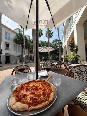 Uma área de refeições ao ar livre com uma pizza na mesa, guarda-sóis e vista para edifícios e palmeiras.