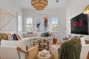 A modern living room with a sectional sofa, a coffee table with snacks, a painting, and a large TV screen displaying the Netflix logo. - Suncerre- 100 Steps to Private Beach & Pool in Carillon Beach (Panama City Beach)
