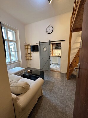 Ein Wohnzimmer mit einer Couch, einem Couchtisch, einem Fernseher an einer Wandablage, einer Uhr und einer Schiebetür, die in eine Küche führt.