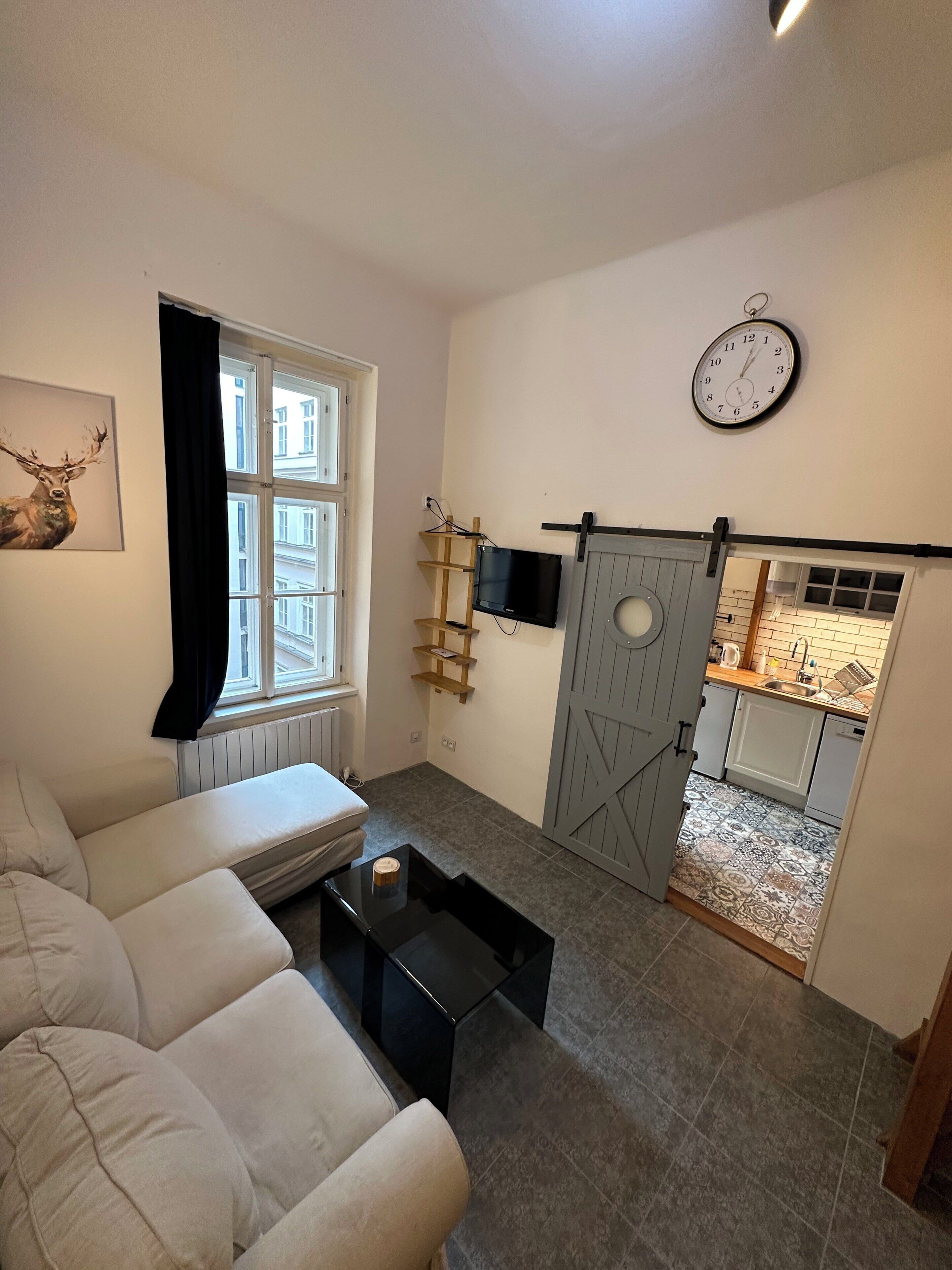 Ein Wohnzimmer mit einer Couch, einem an der Wand montierten Fernseher, einer Uhr und einer Tür zu einer Küche.