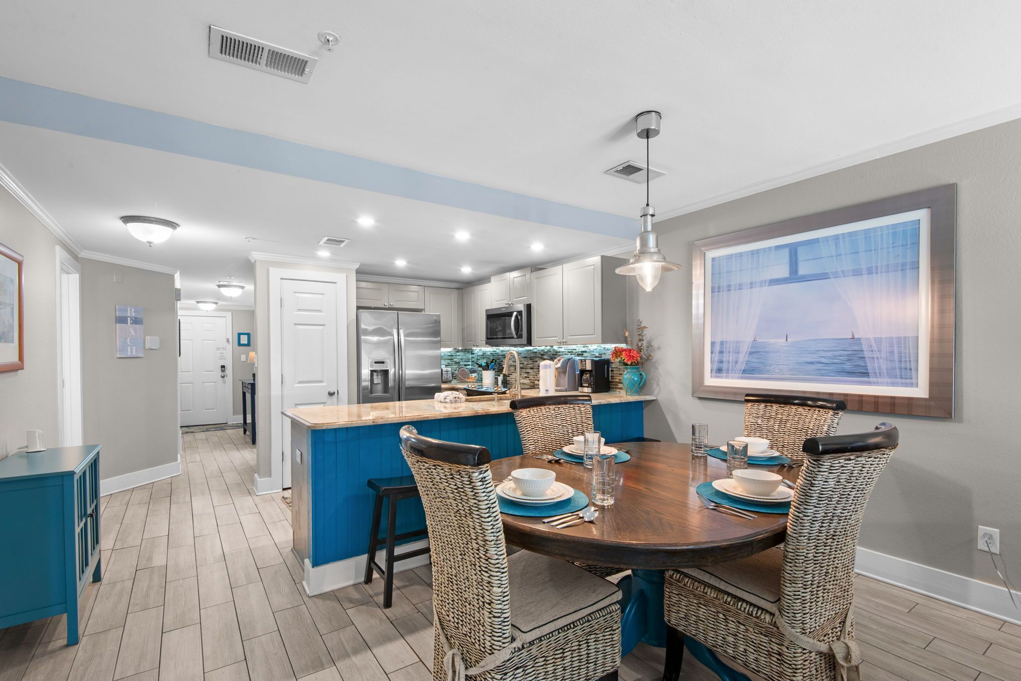 A modern kitchen with a central island, dining area with wicker chairs, and a large window with a view.