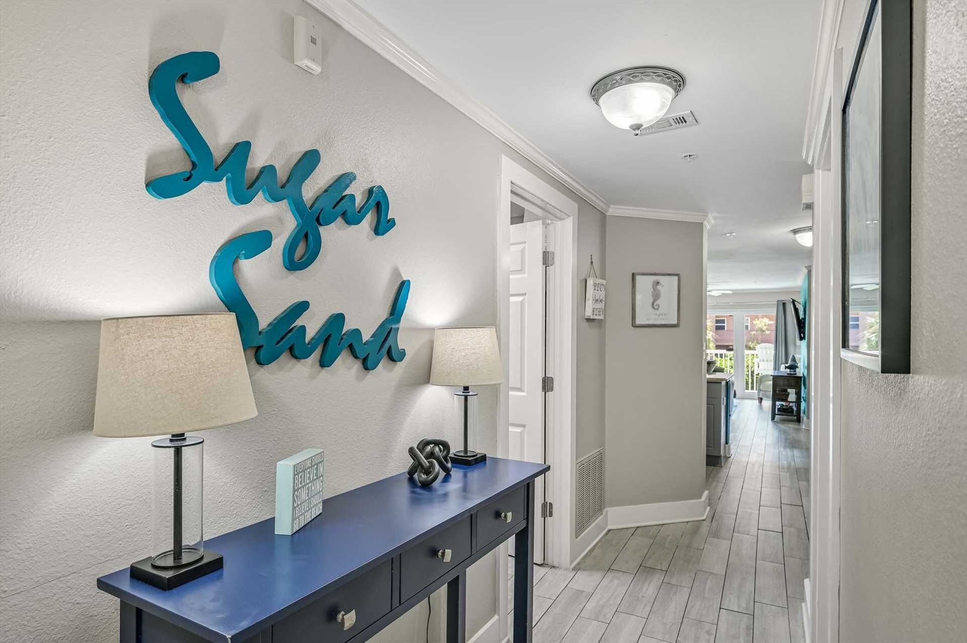 Ein Flur mit einem dekorativen, türkisfarbenen Schild mit der Aufschrift 'Sugar Sand', einem blauen Konsolentisch mit Lampe und einem gerahmten Motivationsspruch.