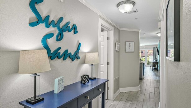 En hall med en dekorativ, turkosfärgad skylt med texten ”Sugar Sand”, ett blått konsolbord med en lampa och en inramad, inspirerande citat.