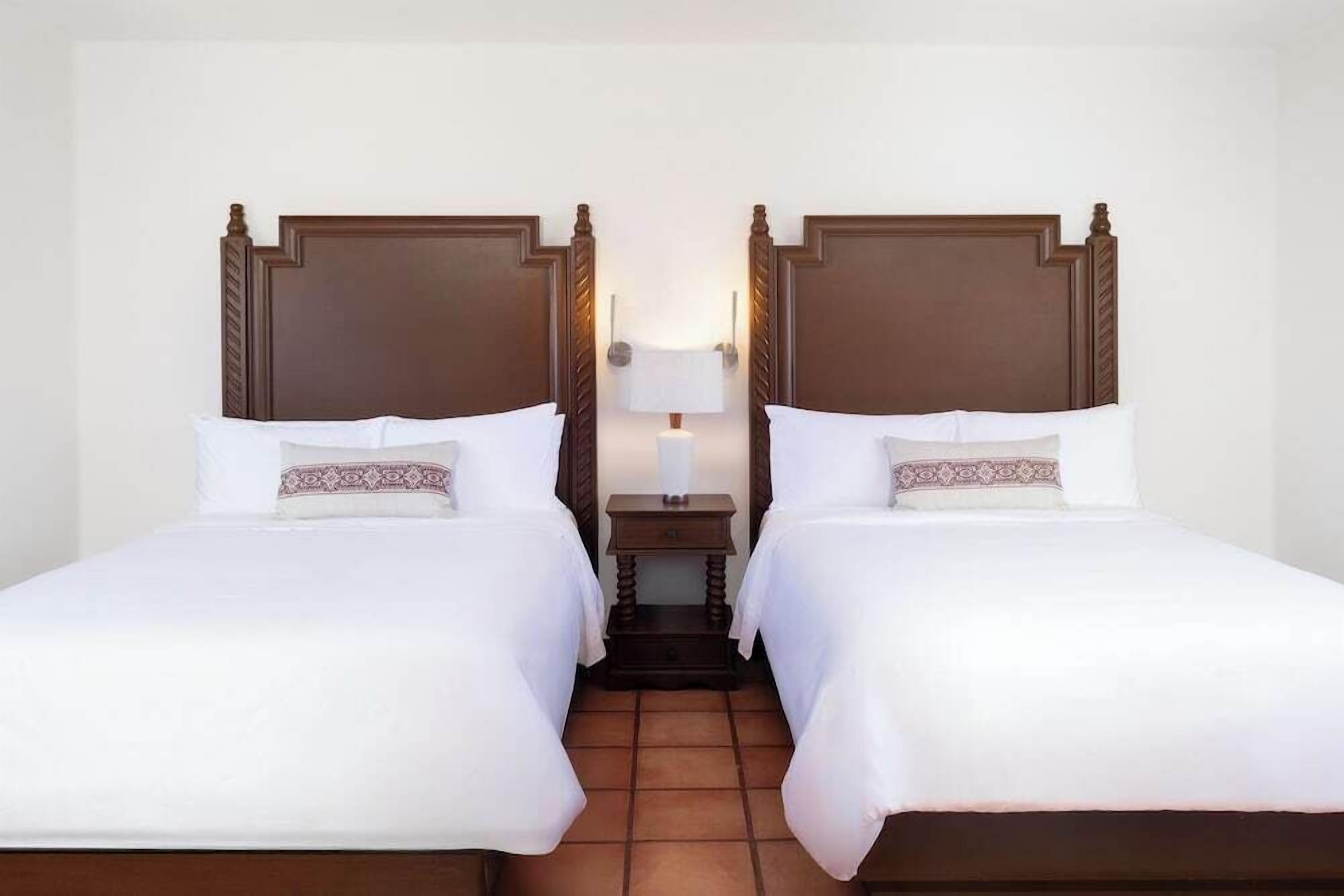 A hotel room with two single beds, a nightstand with a lamp, and a tiled floor.
