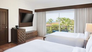 Een hotelkamer met twee bedden, een aan de muur bevestigde televisie, een balkon met uitzicht op palmbomen en een houten dressoir.