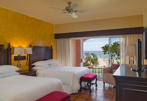 Une chambre d’hôtel avec deux lits, un bureau, une chaise, un ventilateur de plafond et une vue sur la plage.