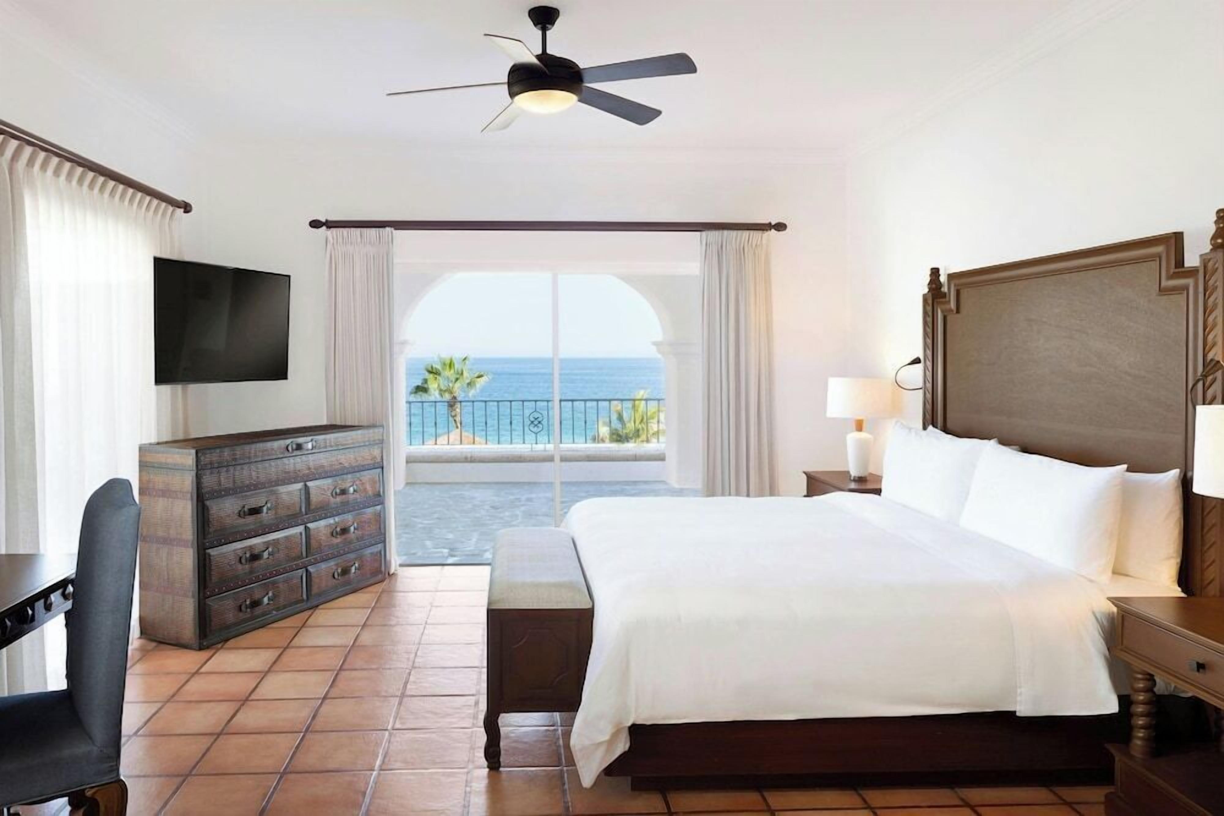 A hotel room with a bed, a desk, a dresser, a TV, a ceiling fan, and a balcony with a view of the sea.