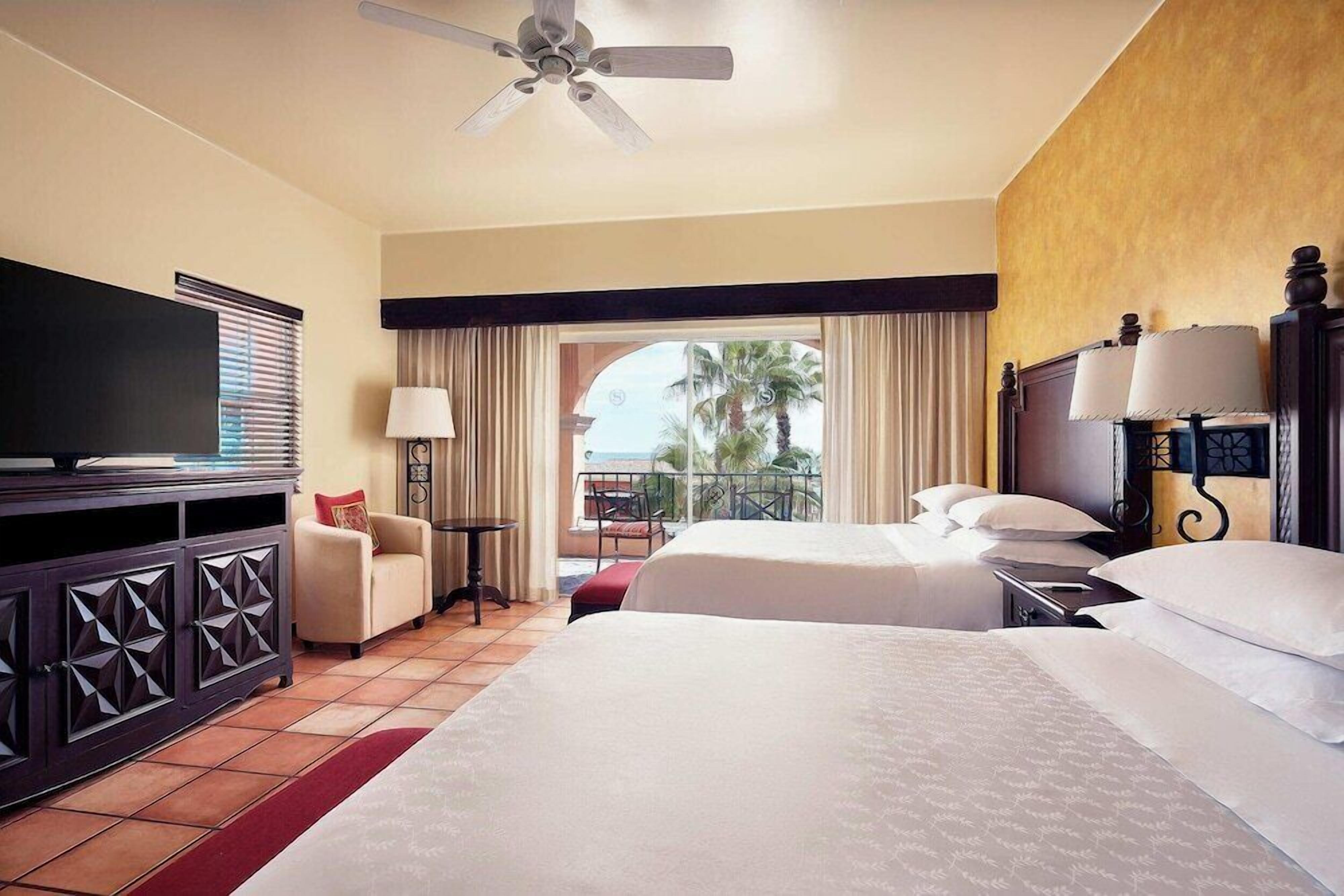 A hotel room with two beds, a television, a desk, and a balcony with a view of palm trees.