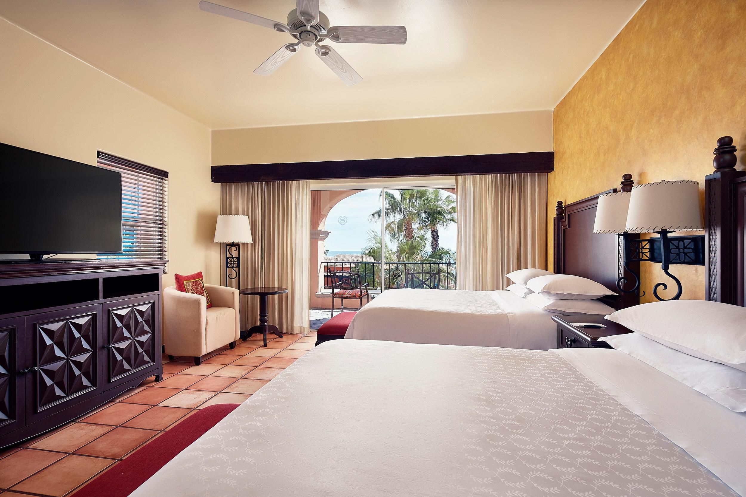 A hotel room with a bed, a TV, a desk, and a balcony with a view of palm trees.