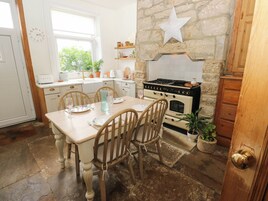 Una cocina con pared de piedra, estufa antigua, sillas de madera y una mesa para dos comensales.