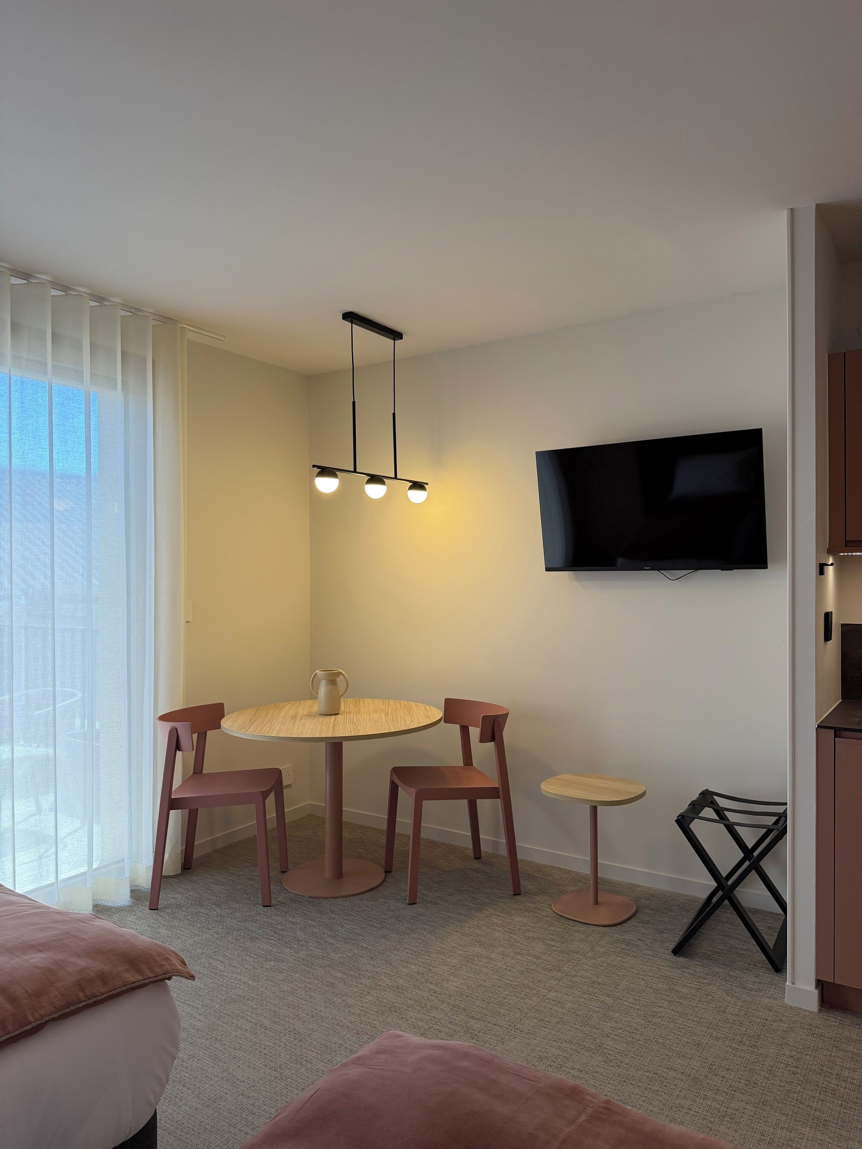 Un salon moderne avec une table de salle à manger ronde, deux chaises, une petite table d’appoint, un téléviseur fixé au mur et une fenêtre ornée de rideaux légers.
