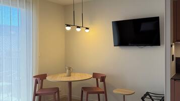 Un salon moderne avec une table de salle à manger ronde, deux chaises, une petite table d’appoint, un téléviseur fixé au mur et une fenêtre ornée de rideaux légers.