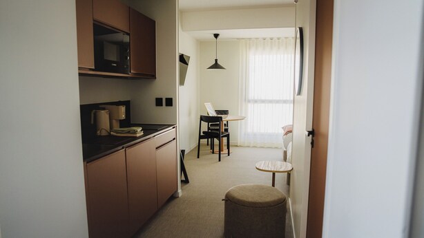 Un couloir étroit, avec une cuisine sur la gauche, un coin repas avec une table et des chaises au centre, et une petite table ronde avec une chaise sur la droite.
