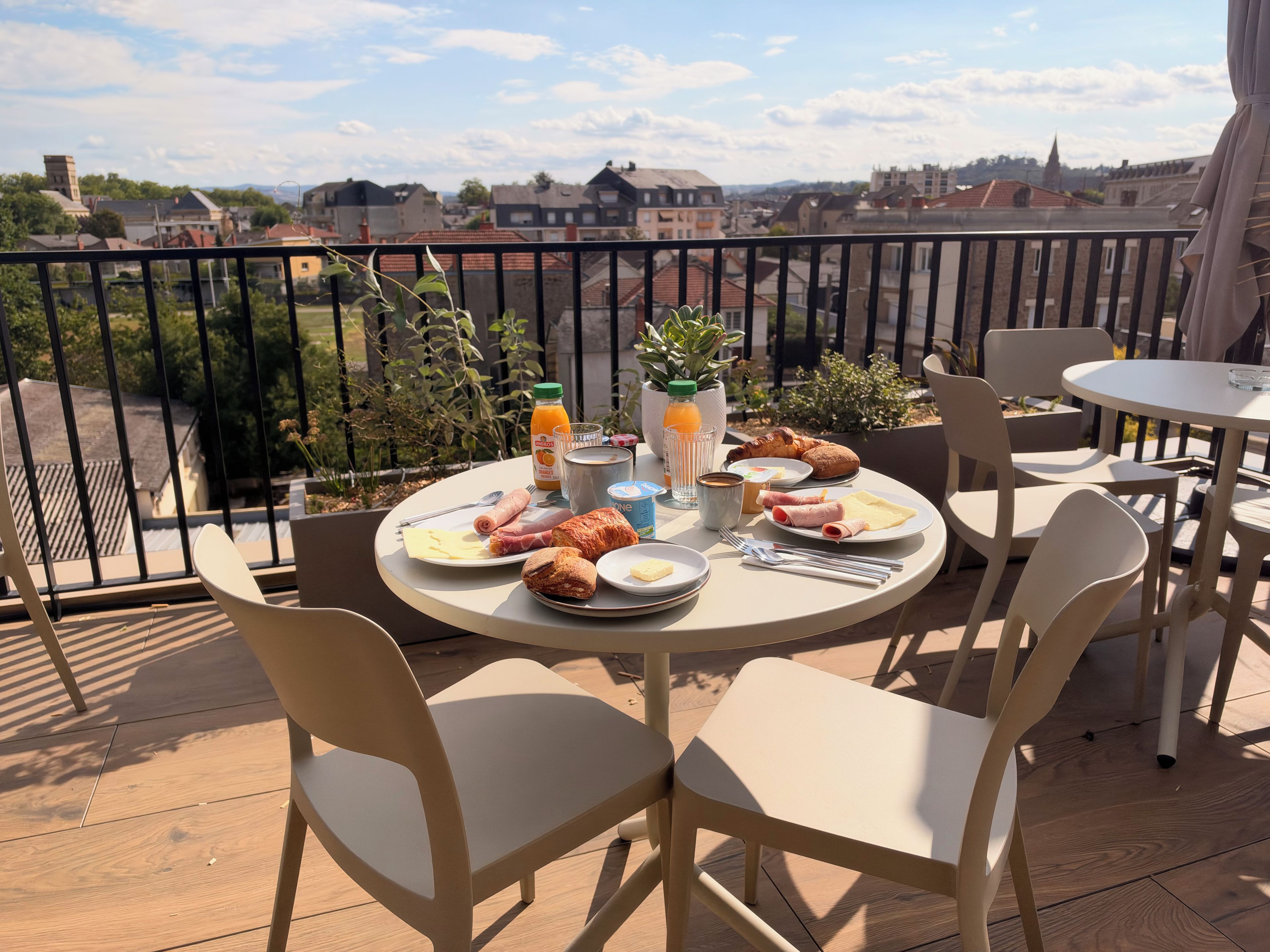A balcony with a table set for breakfast, including croissants, ham, and eggs, with a city view in the background.